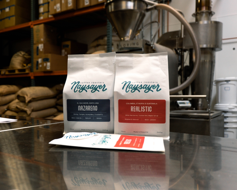 Two bags of Naysayer coffee sitting on a metal table at the Vacera's coffee roasting business with a Loring S15 Falcon roaster in the background. The coffees are labeled Nazareno (single origin) and Realistic (blend).