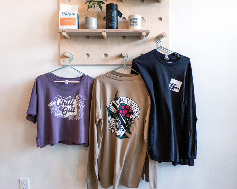 Naysayer Coffee shirts hanging beneath a shelf that features their coffee, a branded tumbler, and a branded mug.