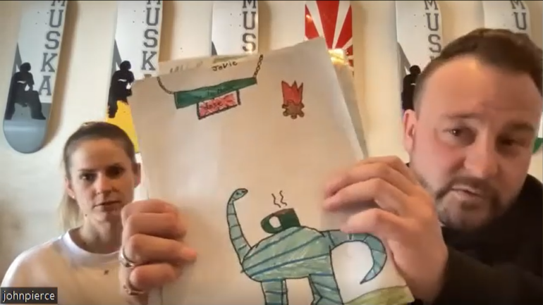 A Zoom screenshot of Lauren Webster and John Pierce. John is showing the camera a child's drawing of a dinosaur with a coffee cup on its back.