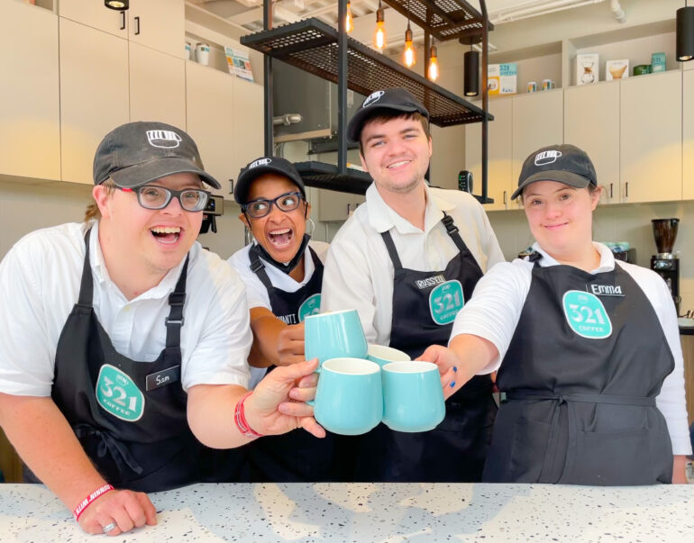 A group of four 321 Coffee employees behind the counter. They are smiling and clinking their turquoise coffee mugs together.