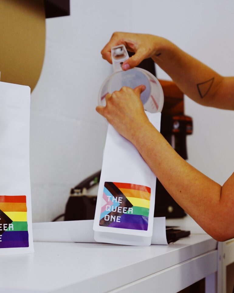 A person's arms are in the shot, a triangle tattoo on their right arm near the elbow. They use a measuring cup to scoop roasted coffee into a white back with a progress flag label that says "The Queer One."