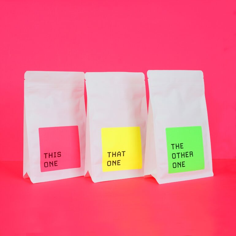 White coffee bags against a neon pink background. Each bag features a neon-colored label that lists This One, That One, and The Other One.