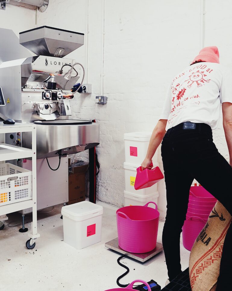 Em Herriot scoops green coffee with bright pink scoops, preparing to roast on the Loring S7 Nighthawk on their left.